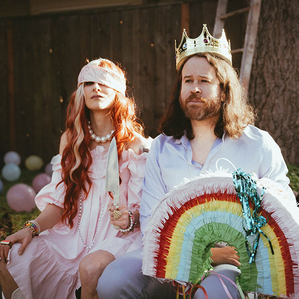 Folk-pop duo Fox and Bones sitting outside; a woman in a pink dress and headband holding a prop knife next to a man wearing a gold crown and holding a rainbow piñata.